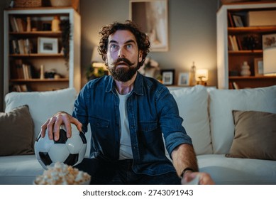 Man feeling anxious while watching a football game on television, holding a soccer ball
