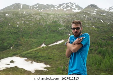 A Man Feeling Cold Because He Is Underdressed Against The Background Of Mountains With Some Snow On Top