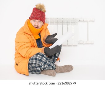 Man Feeling Cold In Hat And Down Jacket Sitting Close To Radiator And Hold High Electricity Gas Bill On White Background. Gas Crisis Concept
