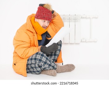 Man Feeling Cold In Hat And Down Jacket Sitting Close To Radiator And Hold High Electricity Gas Bill On White Background. Gas Crisis Concept
