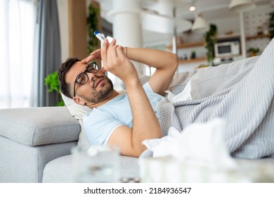 Man Feeling Sick Lying In The Bed And Looking The Thermometer. Sick Man Lying On Sofa Checking His Temperature Under A Blanket At Home In The Living Room