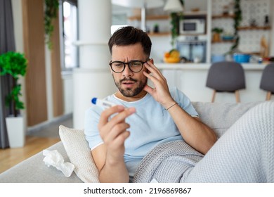 Man Feeling Sick Lying In The Bed And Looking The Thermometer. Sick Man Lying On Sofa Checking His Temperature Under A Blanket At Home In The Living Room