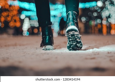 Man Feet In Dark Green Boots, Winter Walking In The Dark Night Time. Shoe Soles Covered With The Snow. Bokeh Lights In The Background Of The Christmas Market