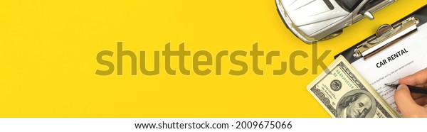 Man\
filling car rental contract. Clipboard on office table with dollars\
and car toy. Yellow background, banner, top view\
