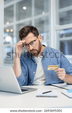 Man finding it difficult to manage debt while holding a credit card and using a laptop for online banking, feeling overwhelmed by financial problems and budget constraints