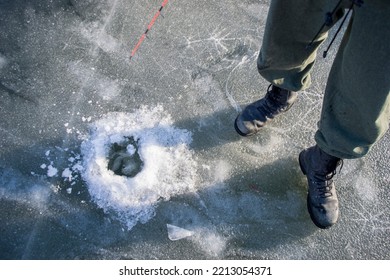 A Man Fisherman In A Jacket, Pants And Boots Stands On A Frozen River Near A Drilled Hole In The Ice And Catches Fish. Body Parts