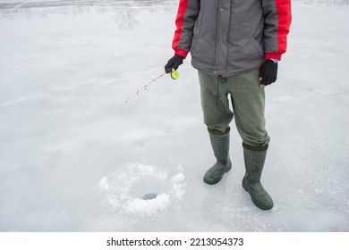 A Man Fisherman In A Jacket, Pants And Boots Stands On A Frozen River Near A Drilled Hole In The Ice And Catches Fish. Body Parts