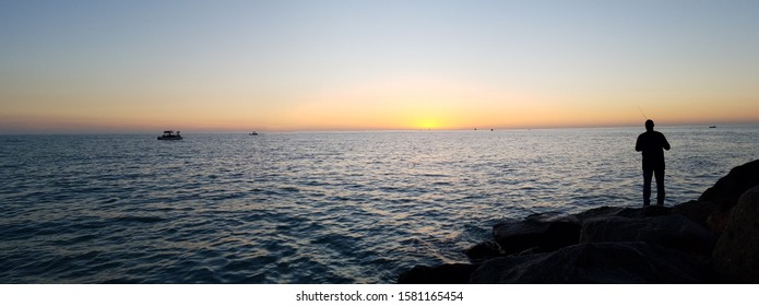 Man fishing along rocky pier amongst ocean sunset