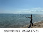 Man fishing on beach at sunset, catching fish rod into calm sea or ocean, outdoor adventure, relaxing vacation, fisherman silhouette, fishing hobby concept. Fisherman for pike, perch, carp.