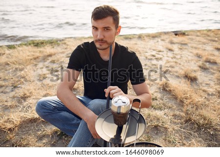 A man is fixing a hookah. He wears a black T-shirt. The man has a beard.