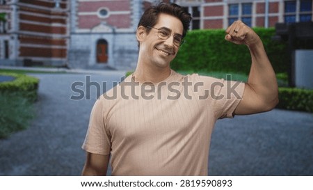 Man flexes bicep and smiles while raising fist in front of a redbrick building entrance, wearing glasses and short sleeve shirt; confidence casual.