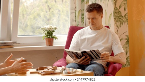 Man flips through a book while sitting in a chair at home. Engrossed in reading, he enjoys a moment of relaxation and leisure in the comfort. Finds solace and enrichment within the pages of the book.