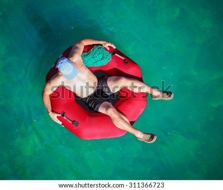 a man floating down a river in a blow up tube with a baseball cap on and shorts on a hot summer day from overhead 