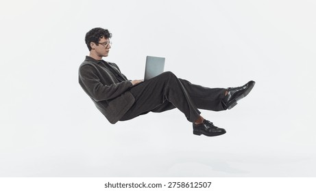 Man floating while using laptop isolated on white background minimal studio shot. Concept of remote work, digital workflow, flexibility, freelance lifestyle, and online productivity