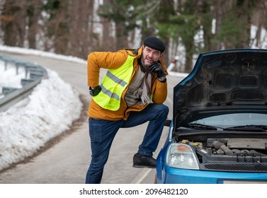 Man With Fluorescent Vest Standing Beside Broken Car With Open Hood Beside Road In Winter Condition With Snow On Grass