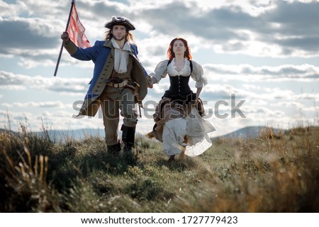 Man in form of officer of War of Independence and girl in historical dress of 18th century. July 4 is US Independence Day. Couple of patriots freedom fighters in outdoor on background cloudy sky
