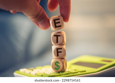 A man forming the acronym ETF for Exchange Traded Fund with wooden dice that are stacked on top of a calculator. A money symbol in the composition.
