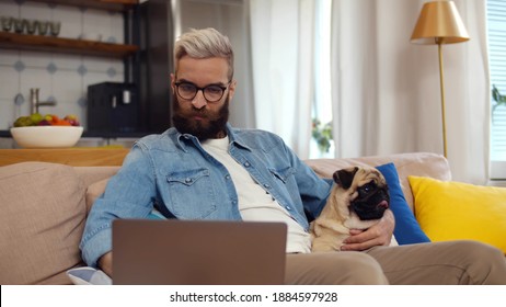 Man Freelancer Working At Home Office With Laptop And Pug Dog On Arms. Hipster Guy In Eyeglasses Relaxing On Couch With Pet And Surfing Internet On Computer At Home