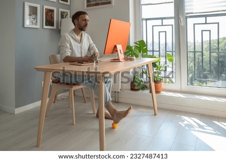 Man IT freelancer working on computer at home doing foot exercise using massage ball for improving blood circulation in legs and spine during long sedentary work. Prevention of leg diseases concept.