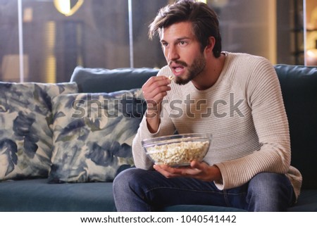 A man in front of the television watches a game of sports sitting on the couch while eating popcorn. Concept of: sports, relaxation, fanfare, betting.