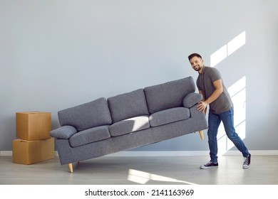 Man Furnishing His New Apartment. Happy Guy Placing Sofa In The Living Room. Strong Boyfriend, Husband Or Father Lifting Soft Comfortable Gray Couch And Smiling. Moving House, Buying Furniture Concept