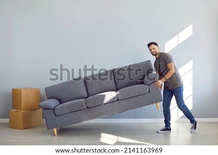 Man furnishing his new apartment. Happy guy placing sofa in the living room. Strong boyfriend, husband or father lifting soft comfortable gray couch and smiling. Moving house, buying furniture concept