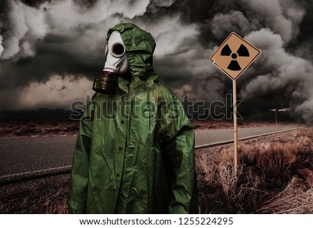 Man in gas mask and cloak for chemical protection on abandoned road with radiation area caution sing under heavy clouds. Nature protection concept