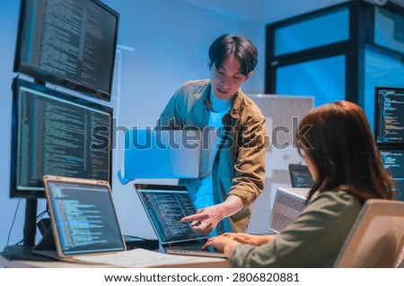 A man gestures toward code displayed on a tablet screen while a seated colleague types on a laptop, surrounded by multiple monitors showing programming code in a blue-lit office.