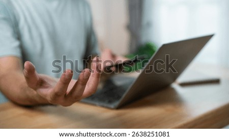 Man gesturing with frustration while using a smartphone and laptop in a cozy workspace, emphasizing communication challenges and technology reliance