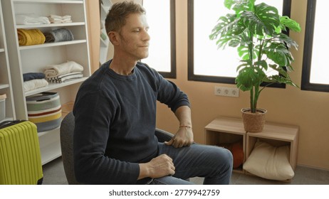 Man gesturing with hands while seated in chair beside potted plant and yellow suitcase in studio; calm reflection.