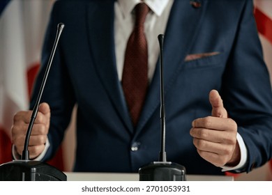 Man gesturing with hands while standing behind podium with microphones, wearing formal suit and tie, delivering speech or addressing audience at event