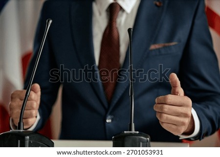 Man gesturing with hands while standing behind podium with microphones, wearing formal suit and tie, delivering speech or addressing audience at event