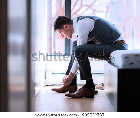 The Man gets his elegant shoes on in his bedroom. Young handsome businessman sitting on bed in hotel room tying his stylish shoes. Groom dressing up with classic, elegant shoes on wedding day