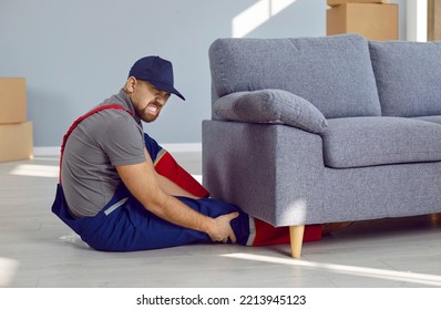 Man Gets Injured While Lifting Heavy Furniture Alone. Professional Handyman Who Got Leg Caught Under Sofa Sitting On Floor And Holding His Broken Knee With Expression Of Pain On Face. Injury Concept