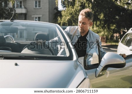 Man getting out of a silver convertible car, holding the steering wheel and door. Arrival home, urban commute, daily routine.