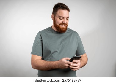 A man with a ginger beard is holding a cell phone in his hand, looking at the screen with a focused expression. He appears to be typing or scrolling on the device.