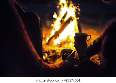 man with the girl sit around the campfire at night with cups
