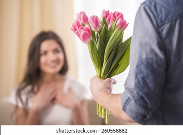 Man Is Giving A Bunch Of Flowers And Happy Woman At Home. Blurred Background.