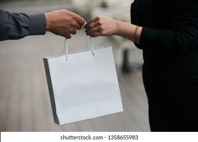 Man Giving Gift In Paper Bag To Woman, Close-up. Gift Giving Between A Man And His Wife. Male Hand Give Shopping Bag To Female Hand