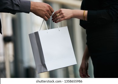 Man Giving Gift In Paper Bag To Woman, Close-up. Gift Giving Between A Man And His Wife. Male Hand Give Shopping Bag To Female Hand