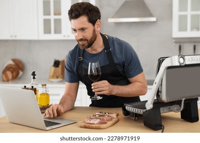 Man with glass of wine making dinner while watching online cooking course via laptop in kitchen - Powered by Shutterstock