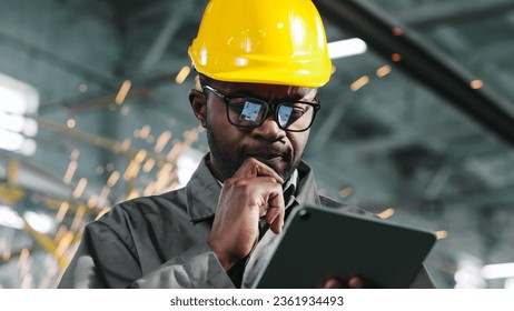 Man in glasses is calculating something on his gadget and is seriously thinking about something. African american manager in special uniform thinks about organization of work in workshop.
