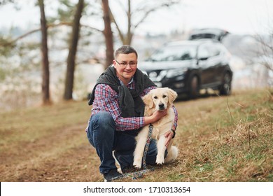 Man In Glasses Have A Walk With His Dog Outdoors In Forest. Modern Black Car Behind.