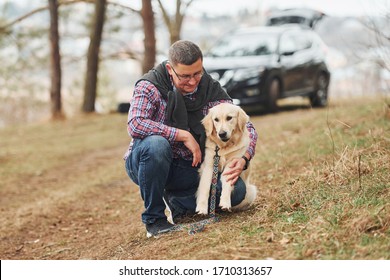 Man In Glasses Have A Walk With His Dog Outdoors In Forest. Modern Black Car Behind.