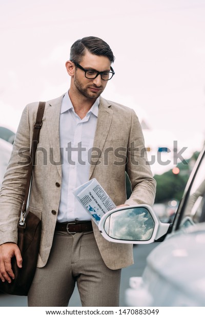 man in glasses holding newspaper and standing near
car on street
