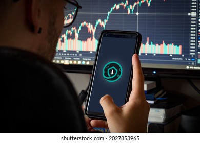 Man With Glasses Holding Phone With Crypto Asset Safemoon On Screen And Defocused Chart Background.