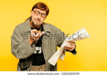 A man in glasses and a jacket scrutinizes a compass while holding an unfolded map, a picture of puzzlement against a yellow backdrop.