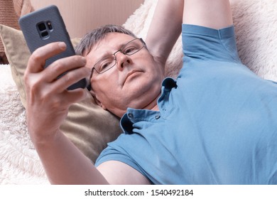 A man with glasses looks at a smartphone while lying in a cozy sofa. Middle-aged businessman checks mail in a smartphone