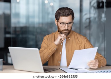 A man in glasses reviews documents while working on his laptop in an office setting.