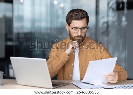 A man in glasses reviews documents while working on his laptop in an office setting.